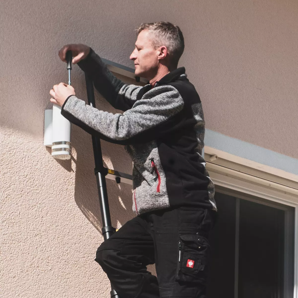 Mann repariert stehend auf der Aluminium-Leiter die Lampe an der Hauswand